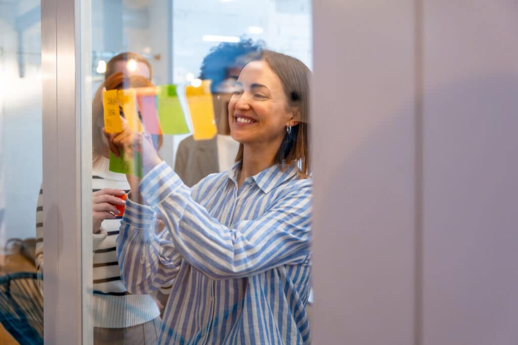 Smiling woman placing colorful sticky notes on a glass wall during a nonprofit board committees planning session.