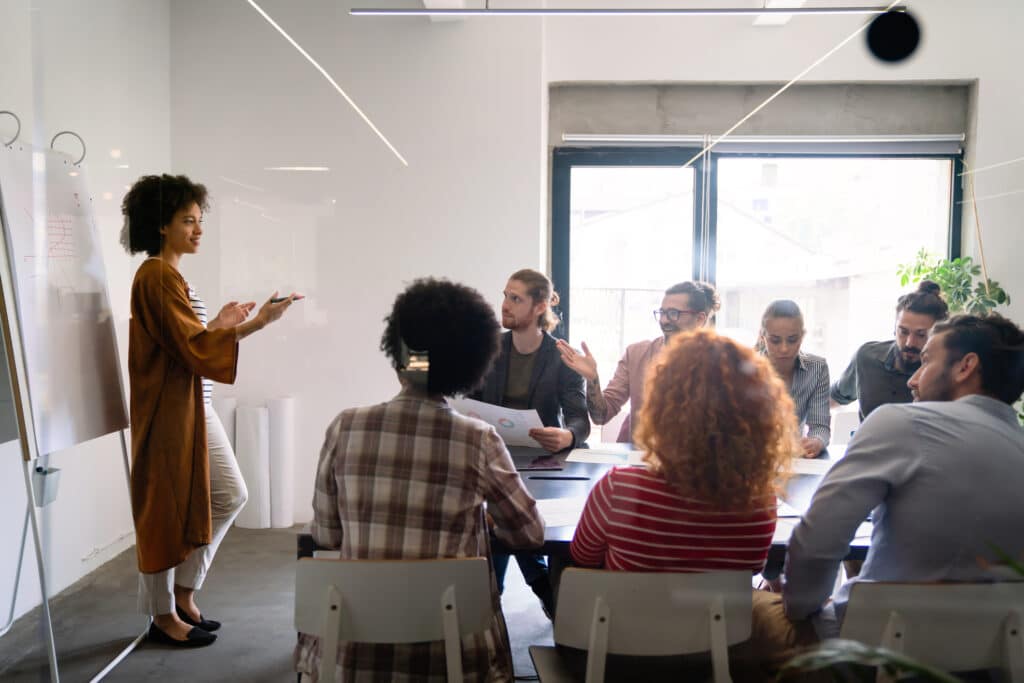 Diverse nonprofit board meeting in a bright conference room: a woman stands at the whiteboard presenting charts to seated colleagues, illustrating the role of board chair not-for-profit governance and facilitation.