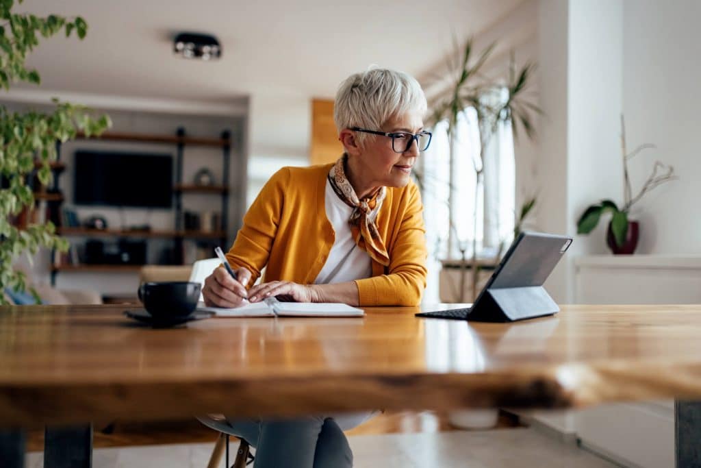 Woman with short gray hair and glasses taking notes while looking at ipad.