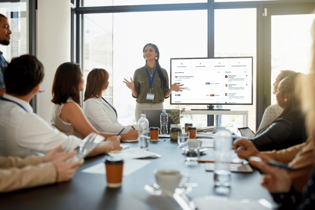 Business woman speaking in front of board at conference table, with monitor in the background.