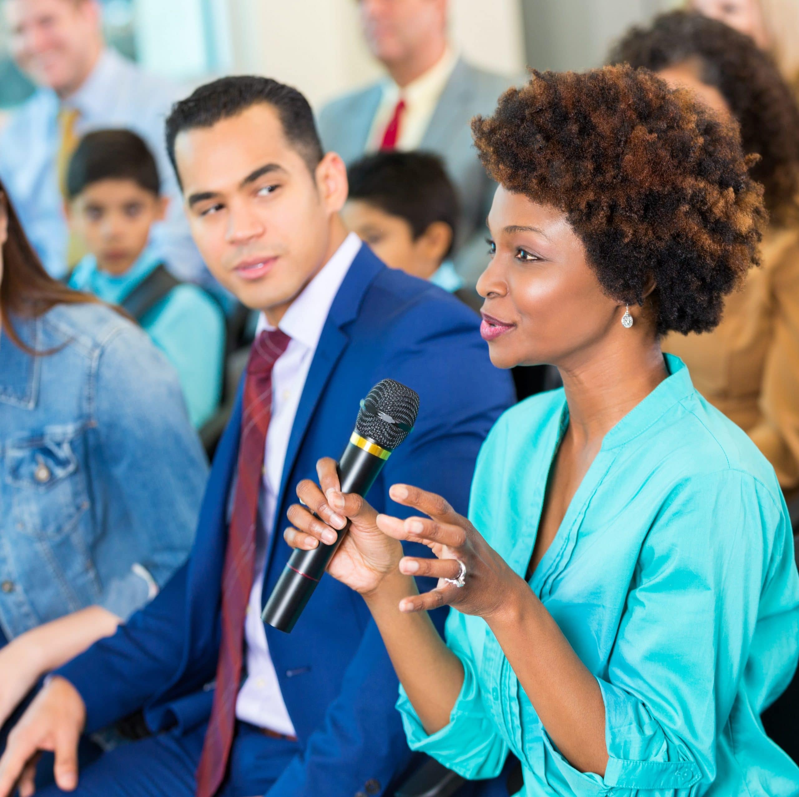 Confident woman asks question during a meeting. A diverse group of people are sitting with her in the audience.