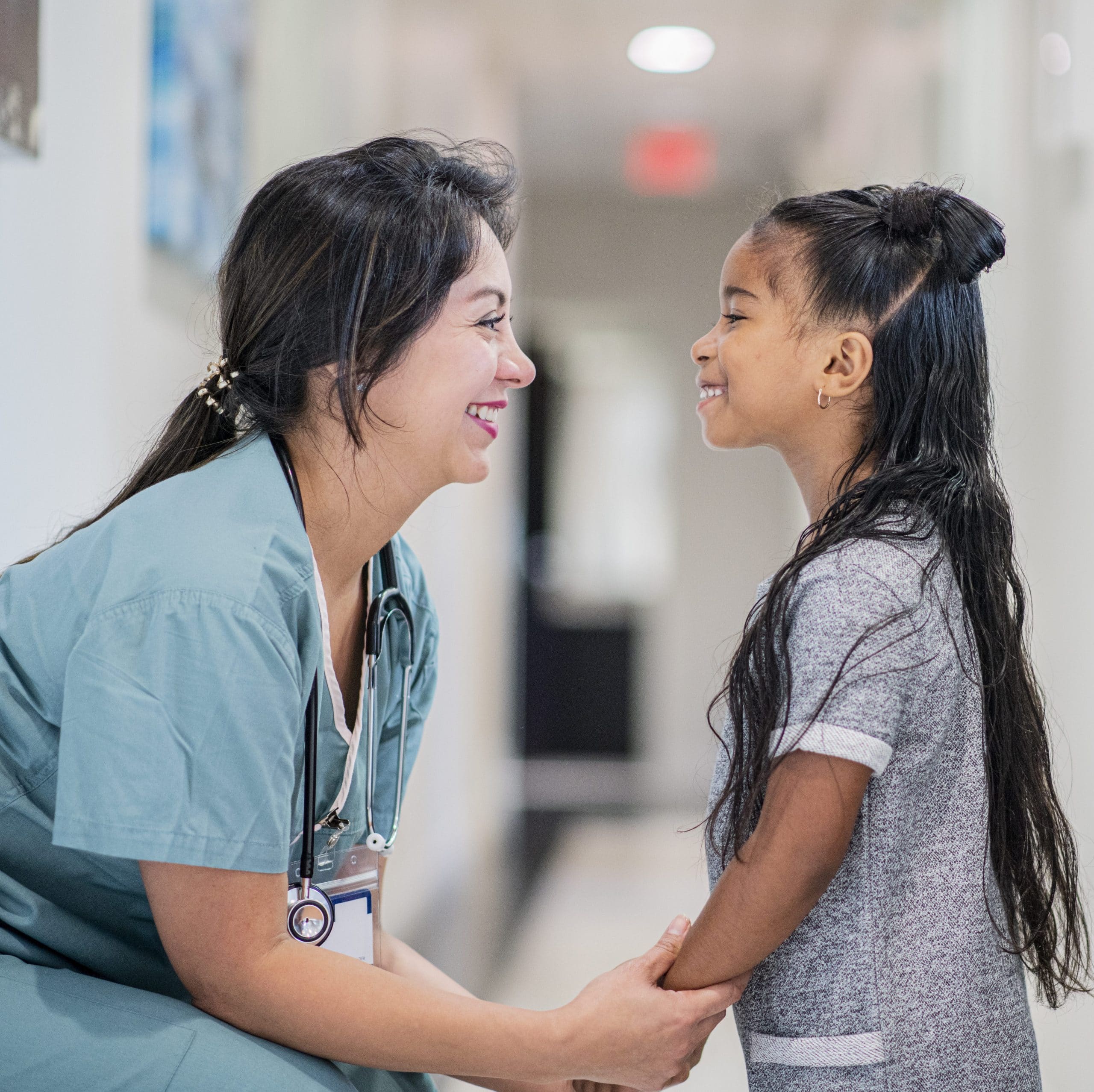 Woman doctor in scrubs, bent down talking to a little girl.