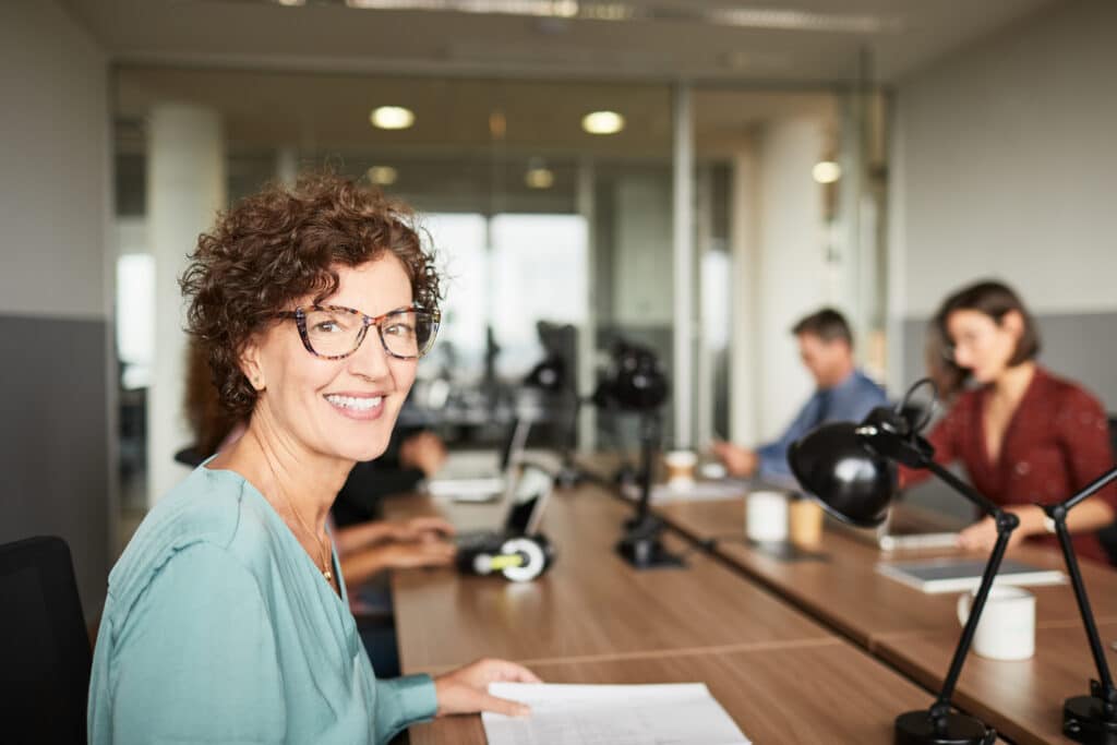 Smiling professional woman in glasses sits at a conference table with colleagues in the background, representing board member positions engaged in collaborative work.