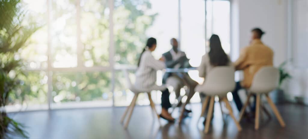 Four diverse professionals sit around a table in a bright, modern conference room with floor-to-ceiling windows, engaged in a blurred discussion—representing different types of nonprofit boards collaborating on strategy and compliance.