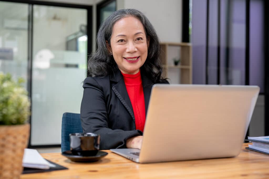 Smiling board member in a red turtleneck and blazer works on her laptop in a bright, modern office—an image that reflects nonprofit board best practices in action.